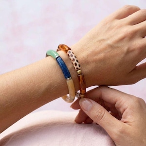 Puede incluir: Dos pulseras elásticas en la muñeca. Una pulsera presenta cuentas cilíndricas en verde claro, azul oscuro y beige. La segunda pulsera consta de cuentas cilíndricas con un patrón de carey marrón y marrón sólido.