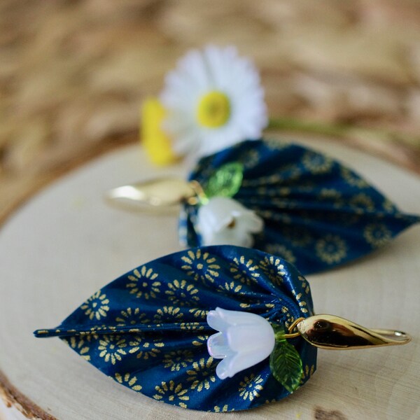 Boucles d'oreilles feuilles origami avec des fleurs dorées sur un fond vert bleu et des petites fleurs blanches