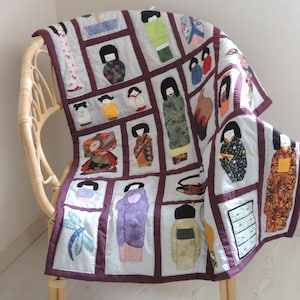 Peut inclure: Une courtepointe en patchwork avec une bordure bordeaux et un fond blanc. La courtepointe présente une variété de poupées japonaises colorées dans différentes poses et tenues. La courtepointe est drapée sur une chaise en osier.