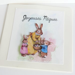 Könnte beinhalten: Aquarell-Osterkarte mit einer Hasenfamilie. Die Mutterhase trägt ein gelbes Kleid und hält einen Korb mit Eiern. Zwei kleinere Hasen tragen rosa und blaue Kleidung. Auf der Karte steht "Joyeuses Pâques".