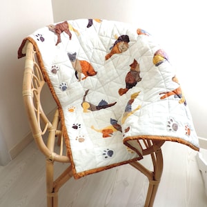 Peut inclure: Une courtepointe blanche avec une bordure brune ornée d'un motif de chats colorés et d'empreintes de pattes. La courtepointe est pliée et drapée sur une chaise en osier.