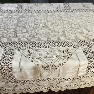 Peut inclure: Une nappe vintage blanc cassé avec des détails de dentelle et de crochet complexes. Le motif présente des motifs floraux et des bordures géométriques. La nappe rectangulaire semble être faite d'un matériau délicat, peut-être du lin.