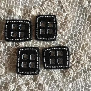 Peut inclure: Quatre boutons carrés noirs avec une finition brillante et une bordure cousue blanche. Chaque bouton comporte quatre ouvertures carrées transparentes. Les boutons sont disposés sur un tissu crème à tissage ouvert.