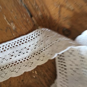 Peut inclure: Gros plan d'une bordure en dentelle blanche crochetée avec un bord festonné. La dentelle présente un motif ajouré avec de petits trous espacés. La bordure est sur une surface en bois.