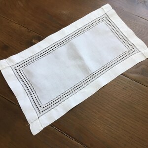 Può includere: Un sottopiatto rettangolare in lino bianco con un bordo decorativo di piccoli fori distanziati. Il sottopiatto è appoggiato su una superficie di legno, evidenziando il suo design pulito. I bordi sono accuratamente orlati.