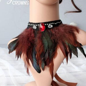 May include: A black choker necklace with a red jewel and clear rhinestones, adorned with brown and green feathers.