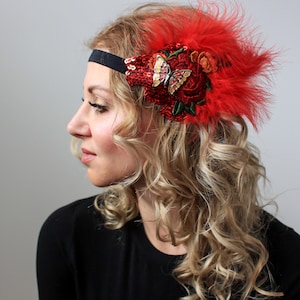 May include: A red flapper headband with a black band. The headband features red feathers, sequins, and embroidered flowers. A gold and cream butterfly is centered on the headband. The accessory is designed for special occasions.