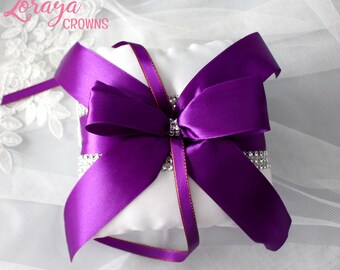 May include: A white ring pillow with a large purple satin bow and a rhinestone embellishment. The pillow is on a white lace background.