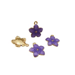Może przedstawiać: Cztery zawieszki w kształcie kwiatów. Trzy są fioletowe ze złotym obramowaniem, a jedna jest złota. Fioletowe zawieszki mają małą pętelkę u góry do zawieszenia. Złota zawieszka ma kształt liścia.