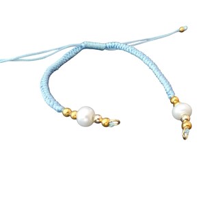 Puede incluir: Una pulsera de macramé azul claro con una perla central y cuentas doradas. La pulsera ajustable presenta un cierre de nudo corredizo y detalles dorados. La pulsera es una pieza de joyería delicada.