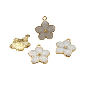 Może przedstawiać: Cztery zawieszki w kształcie kwiatów. Trzy są białe ze złotymi obwódkami i środkami, a jedna jest złota. Każda zawieszka ma pętelkę do mocowania do biżuterii. Zawieszki są do rękodzieła.