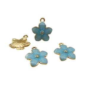 Może przedstawiać: Cztery zawieszki w kształcie kwiatów. Trzy są jasnoniebieskie ze złotymi krawędziami i środkami. Jeden jest w kolorze złotym. Każda zawieszka ma małą pętlę do mocowania do biżuterii lub innych rękodzieł. Zawieszki są na białym tle.