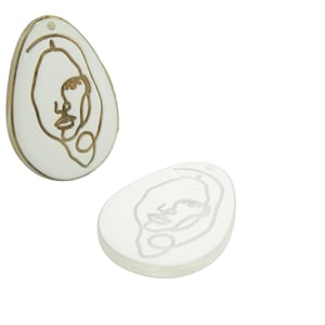 Peut inclure: Deux ornements en céramique blanche en forme d'œuf avec un dessin au trait doré d'un visage sur chacun.