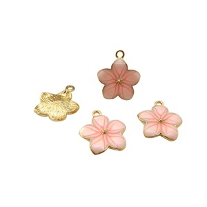 Może przedstawiać: Cztery zawieszki w kształcie kwiatów. Trzy zawieszki są różowe ze złotymi krawędziami i małą złotą pętelką na górze. Jedna zawieszka jest złota z teksturowaną powierzchnią. Wszystkie zawieszki są na białym tle.