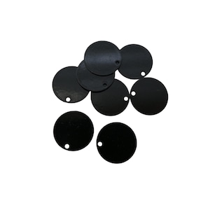 Peut inclure: Un ensemble de sept disques noirs plats et circulaires disposés en groupe. Chaque disque présente un petit trou circulaire percé près du bord extérieur, conçu pour la fixation ou l'enfilage. Les surfaces sont lisses et mates.