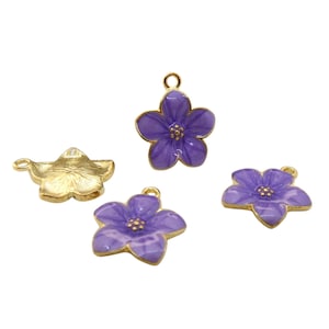 Może przedstawiać: Cztery zawieszki w kształcie kwiatów. Trzy są fioletowe ze złotymi obwódkami i środkami, a jedna jest złota. Każda zawieszka ma pętelkę do mocowania do biżuterii lub innych prac rzemieślniczych. Zawieszki są na białym tle.