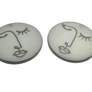 Peut inclure: Deux pendentifs ronds blanc cassé avec un design de visage minimaliste. Chaque pendentif présente un contour noir d'un visage avec les yeux fermés et une bouche. Les pendentifs ont une bordure gris foncé et un petit trou en haut.