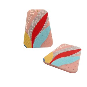 Puede incluir: Un par de pendientes triangulares de color rosa con un diseño abstracto colorido. Los pendientes presentan detalles en rojo, azul, amarillo y blanco.