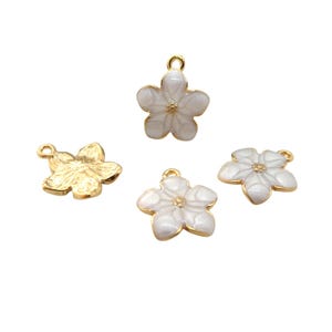 Może przedstawiać: Cztery zawieszki w kształcie kwiatów. Trzy są białe ze złotymi krawędziami i środkami, a jedna jest złota. Każda zawieszka ma pętlę do mocowania do biżuterii. Zawieszki są na białym tle.