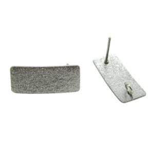 May include: Two silver-colored rectangular stud earrings with a textured surface. One earring is lying flat, while the other is angled, showing the post and earring back. The earrings are set against a white background.