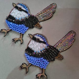 May include: Two embroidered bird patches. Each bird features a blue, white, and black beaded body with iridescent wings and blue sequin accents. The birds have gold-colored feet and are set against a neutral background.