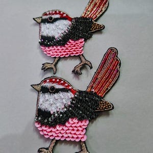 May include: Two embroidered bird patches. Each bird has a red and white head, black sequined wings, and a pink sequined belly. The tail feathers are red and gold. The birds have brown embroidered legs and are set against a gray background.