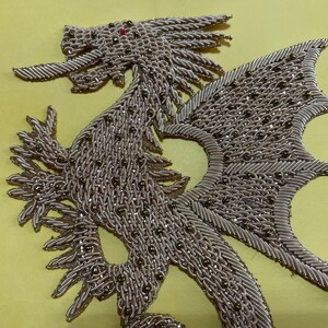 Op de afbeelding: Een goudkleurige drakenapplicatie met een parelmoerontwerp. De draak heeft een lange staart, vleugels en een woeste uitdrukking.