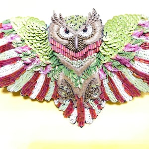May include: An owl-shaped decorative piece with intricate beadwork and sequin details. The owl features a brown and tan body, red eyes, and wings with green, white, and burgundy accents. The wings are spread, creating a symmetrical design.