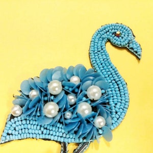 May include: A blue beaded flamingo with white pearl accents. The flamingo is made of small blue beads and has a large, round body with a long, curved neck. The flamingo is decorated with white pearls that are scattered throughout the body and neck.