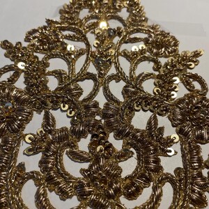 May include: Gold embroidered applique with intricate floral designs and sequins. This decorative embellishment features a delicate, ornate pattern.