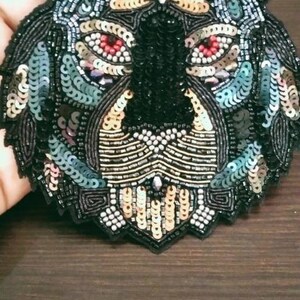 May include: A black, green, and gold sequined tiger face applique. The tiger has black eyes with red accents and a black nose.
