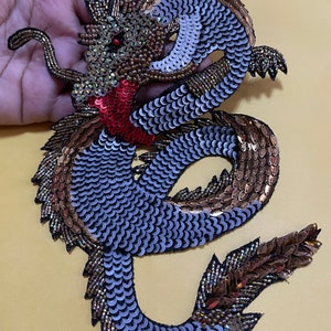 May include: A large, detailed, sequined dragon applique in shades of gold, silver, and purple. The dragon is curled up with its head raised and its tail curled around its body.