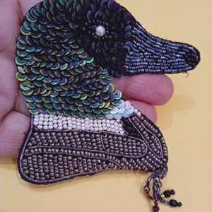 May include: A beaded and sequined brooch in the shape of a duck's head. The duck has a black eye with a white bead in the center, a black beak, and iridescent green and blue feathers. The duck's neck is beaded in brown and white.