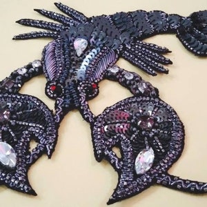 Può includere: Un'applicazione di aragosta in paillettes nere con occhi rossi e accenti iridescenti. L'aragosta è in posizione arrotolata con le chele alzate.