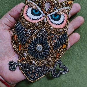 Op de afbeelding: Een gedetailleerde uil-applicatie, rijkelijk versierd met gouden, zilveren en zwarte kralen. De uil heeft blauwe ogen en roze accenten. Geschikt voor het verfraaien van kleding of accessoires.