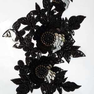 Può includere: Applique floreale nera con perline e paillettes. L'applique presenta tre grandi fiori con una perla nera centrale e paillettes argentate. I fiori sono circondati da perline e paillettes nere più piccole.