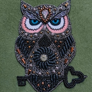 Op de afbeelding: Geborduurde uil patch met kralen. De uil heeft blauwe ogen, roze en witte details en een zwart lichaam met een bloemmotief. Het is versierd met zilveren, gouden en oranje kralen, wat een textuur geeft. Groene achtergrond.