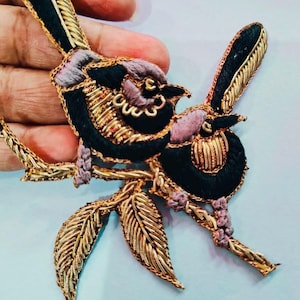 May include: Embroidered bird applique featuring two birds perched on a gold-colored branch with leaves. The birds have black and lavender bodies with gold accents. The leaves and branch are also gold.
