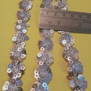 May include: Three strands of silver and white sequined trim with a floral design. The trim is approximately 1 inch wide.