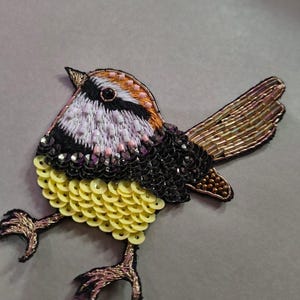 May include: Embroidered bird patch with a detailed design. The bird features a black, white, and orange head, a yellow sequined belly, and a brown beaded tail. The bird's legs are made of gold thread.