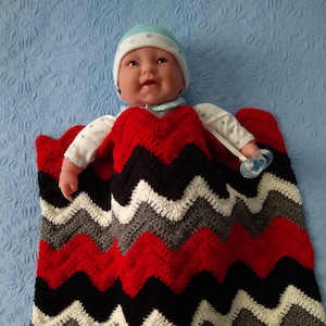 May include: A baby doll wearing a blue hat and a red, black, white, and gray chevron patterned crocheted blanket. The doll is holding a blue pacifier.