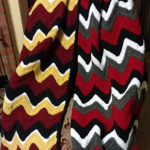 May include: A crocheted chevron blanket in red, black, white, and gold. The blanket is folded in half and draped over a chair.