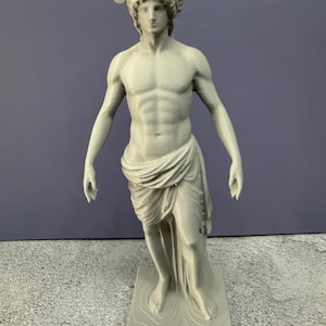 May include: A gray statue of a man with wings on his head, reminiscent of classical Greek or Roman art. The statue is standing on a rectangular base and is draped in fabric.