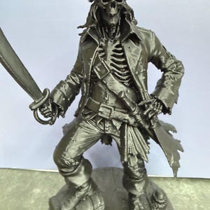May include: A black monochromatic figurine of a skeleton pirate wearing a tattered long coat and a tricorn hat. The figure stands on a chest, holding a sword in one hand, with visible rib cage details and a belt with a sheathed dagger at the waist.