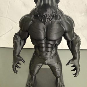 May include: A black 3D-printed figurine of a werewolf. The figure has a muscular physique, with a bear-like head and clawed hands and feet. It is wearing jeans and has a menacing expression.