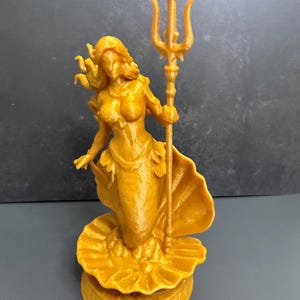 May include: A golden-colored figurine of a mermaid holding a trident. The mermaid is standing on a seashell base. The figurine is approximately 6 inches tall.