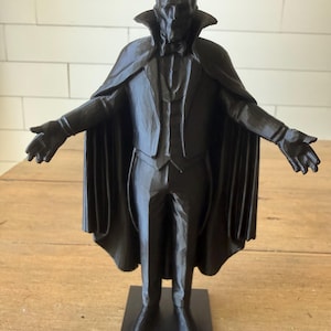 May include: A black statue of a figure in a suit, cape, and bow tie, with arms outstretched. The figure has a bald head and a stern expression. The statue is on a black rectangular base.