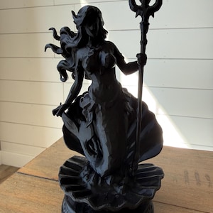 May include: A black sculpture of a mermaid holding a trident. The mermaid has long flowing hair and a fishtail. The sculpture is on a decorative base with a seashell design. The statue is made of a dark material and is displayed on a wooden table.