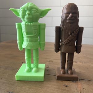May include: Two plastic nutcrackers: one green Yoda figure and one brown Chewbacca figure. Both figures are standing on square bases. The Yoda figure has large ears and is wearing a tunic. The Chewbacca figure has a bandolier.