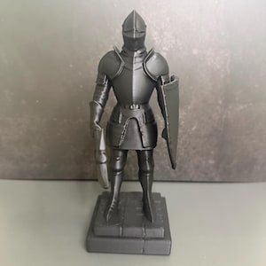 May include: A gray figurine of a knight in full armor, holding a sword and shield. The knight stands on a rectangular base. The armor has detailed features, including a helmet, shoulder guards, and leg protection. The overall design is a medieval theme.